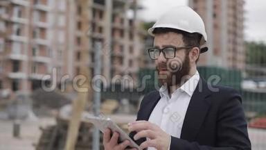年轻的胡子建筑师在工地用平板电脑拍照。 他穿着商务服。